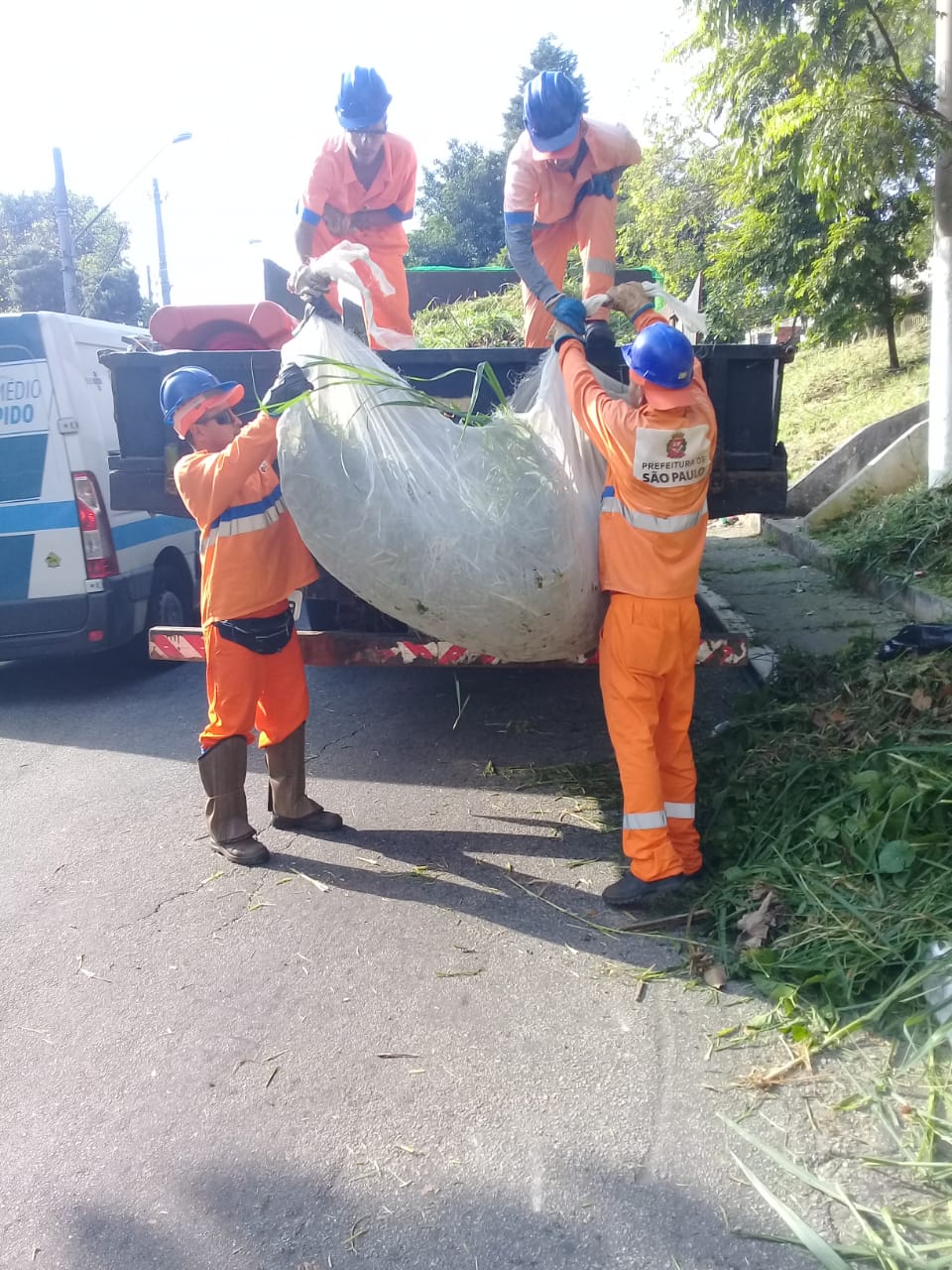 Dois trabalhadores de macacão laranja erguem pacote com mato cortado, para entregar a outros dois que estão em cima do caminhão.